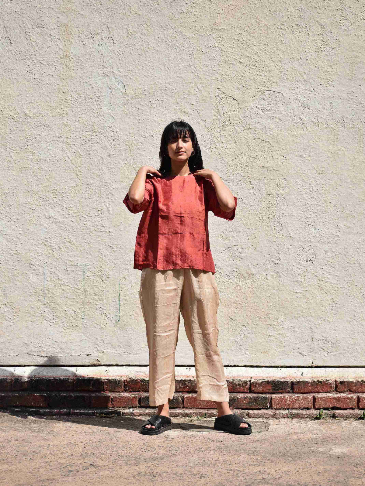 Person wearing a red top and beige pants standing against a textured wall.