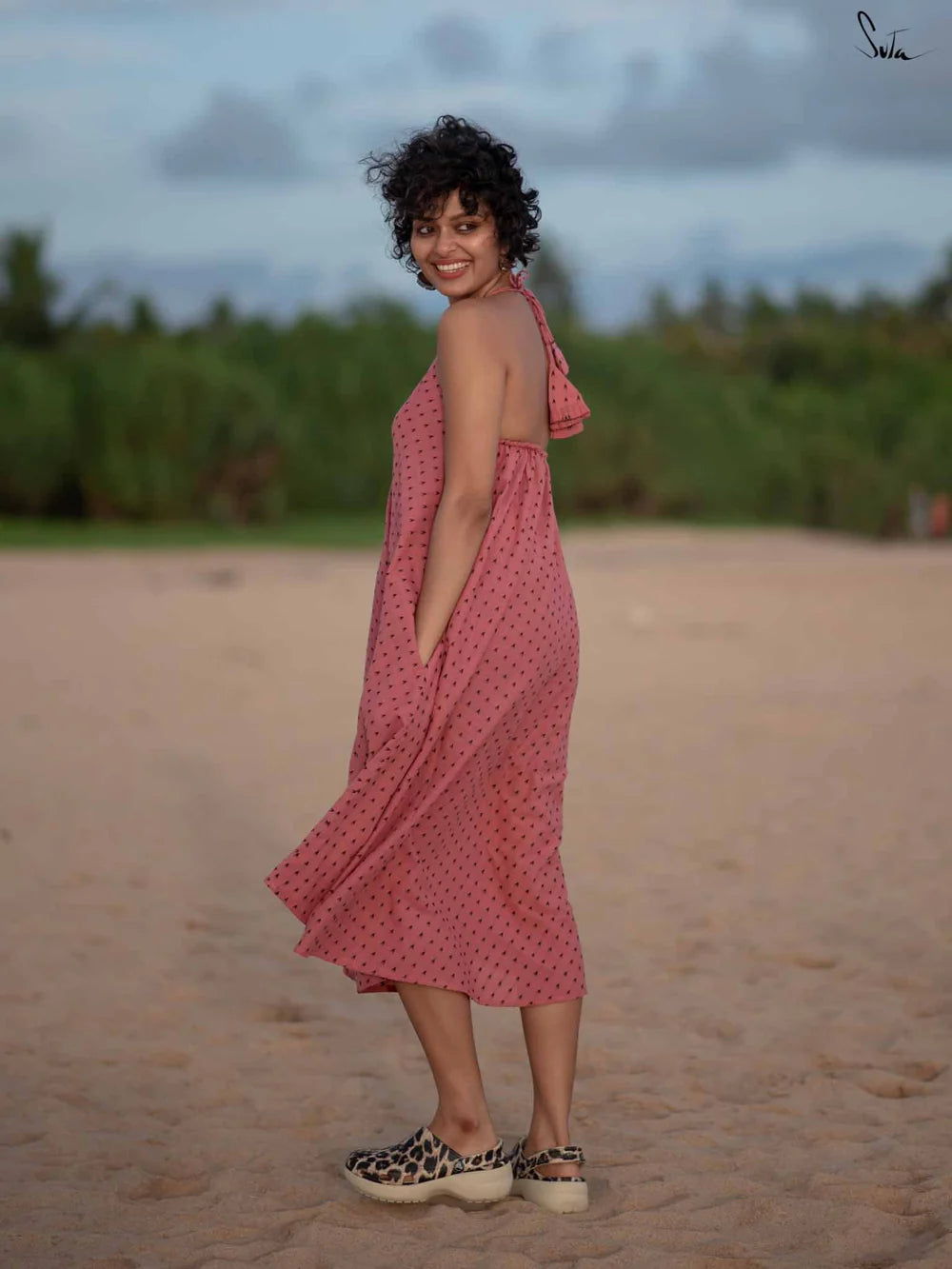 Woman in a pink dress standing on a sandy beach with greenery in the background
