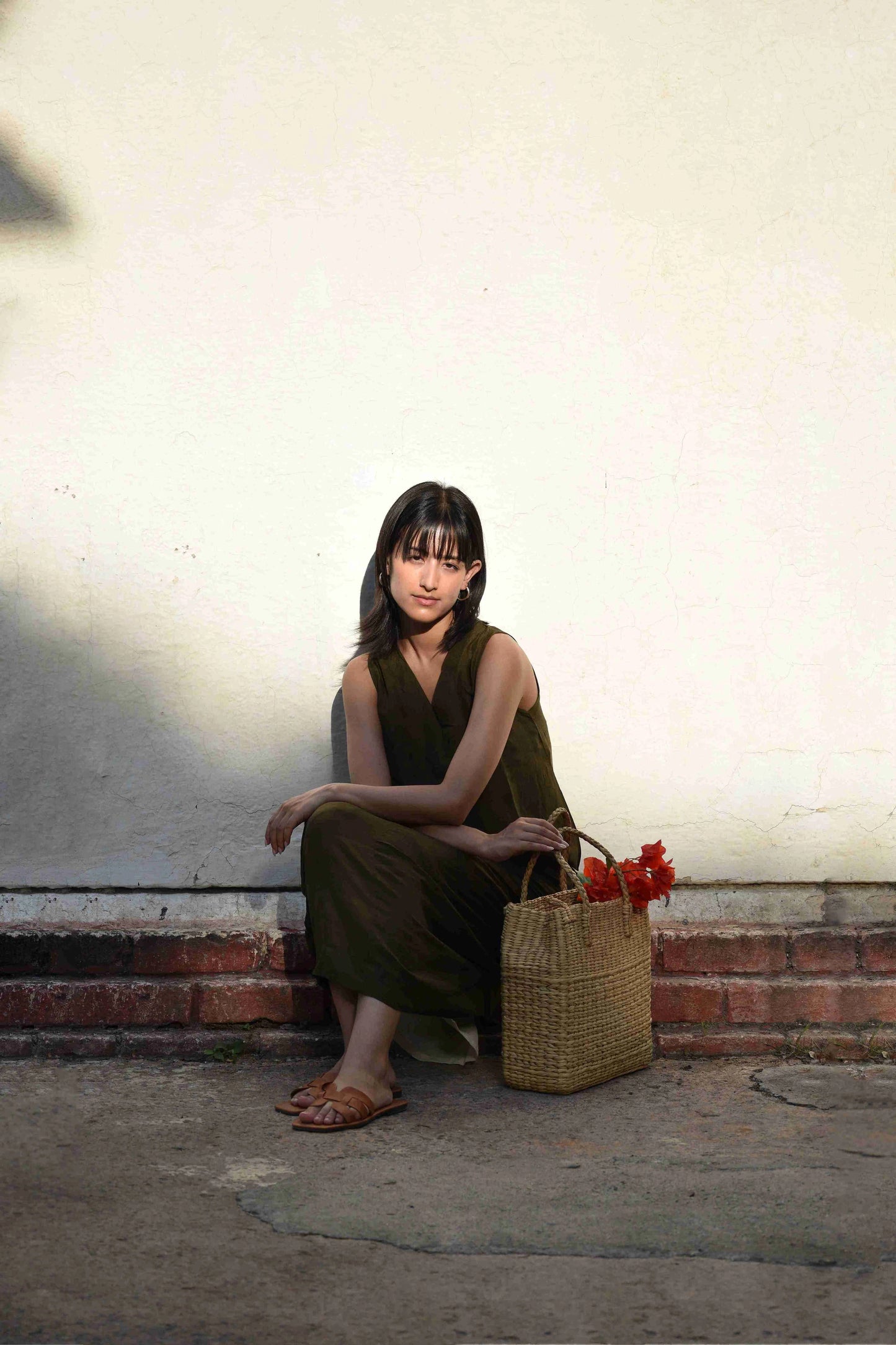 Woman sitting on a step holding a woven bag with flowers against a plain wall.