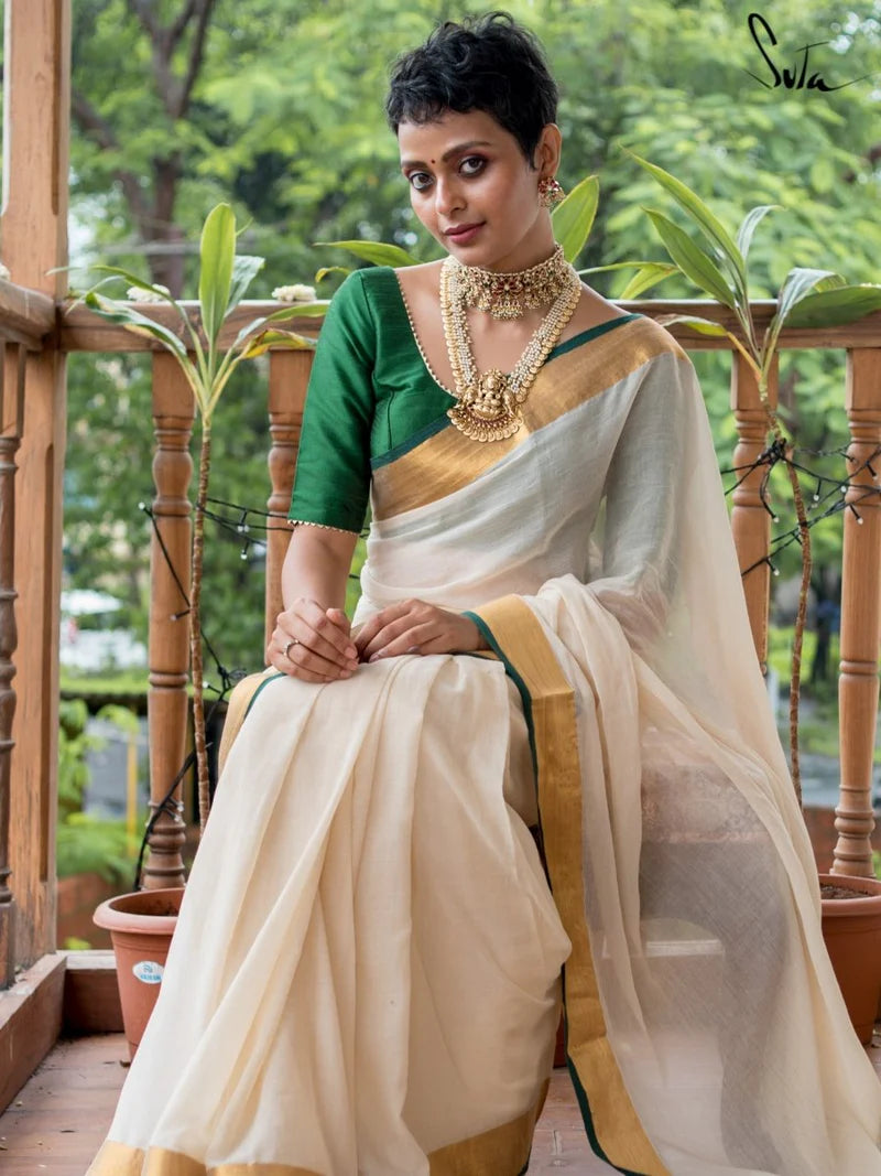 Woman in a green and white saree with gold accents sitting on a wooden deck with plants in the background.