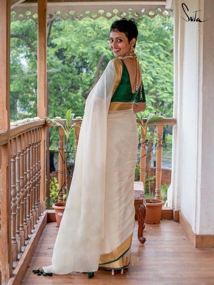 Woman in a green blouse and white saree standing on a wooden porch with greenery in the background