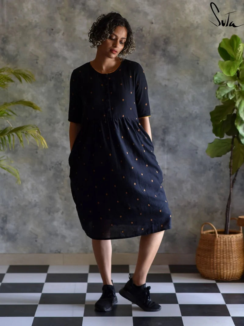 Woman wearing a black dress with gold patterns against a textured wall with plants.